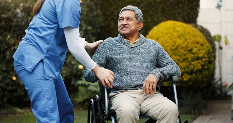 Older adult uses a tablet while a caregiver reviews a digital care checklist in a bright assisted living room.